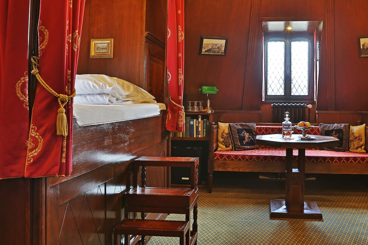 A room with a bed, a round table with a bowl of fruit, a bookshelf, and a window with curtains.