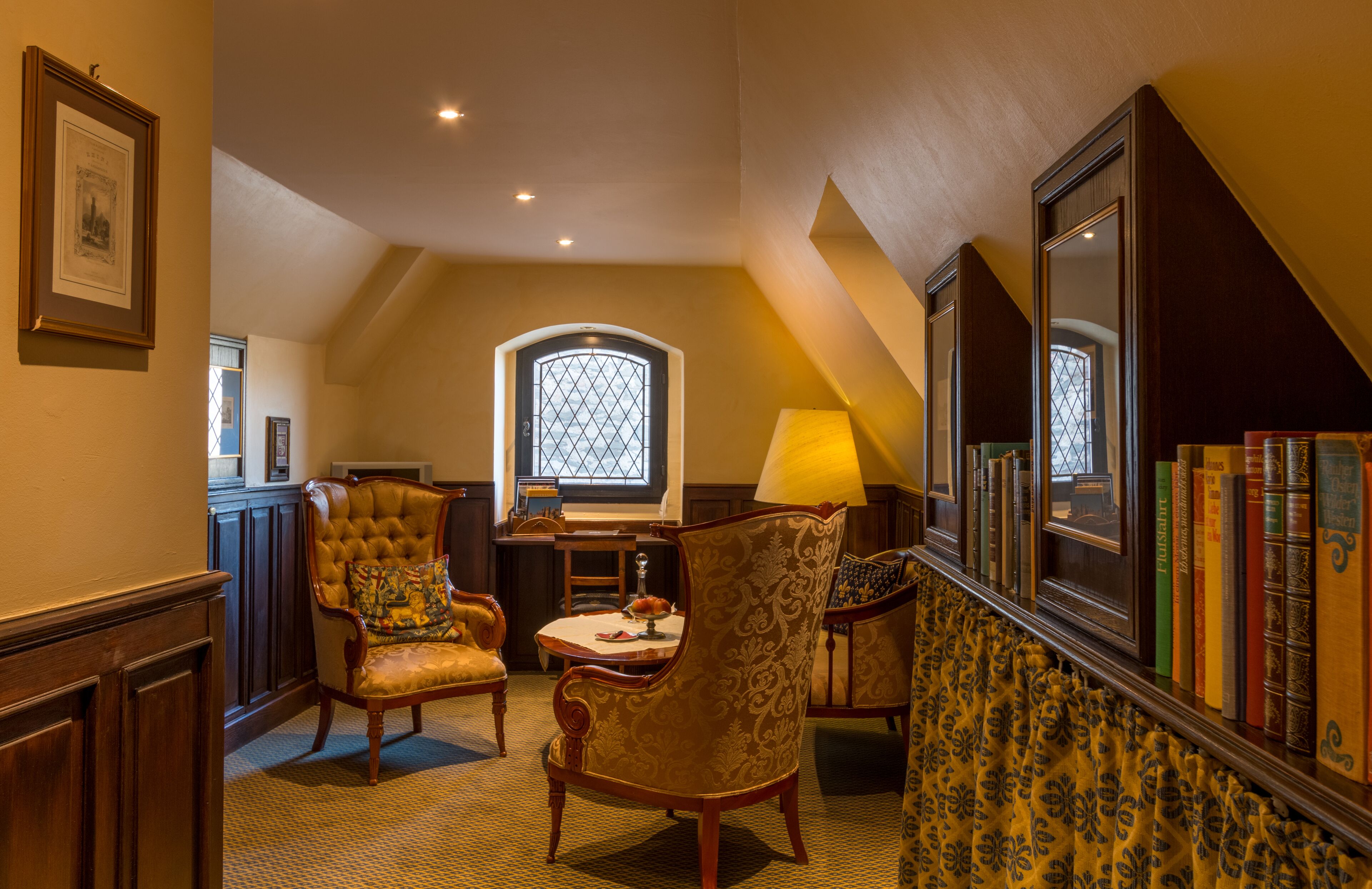 Une pièce avec un plafond haut, une fenêtre ornée d’un motif en losange, une table avec une lampe et une bibliothèque remplie de livres.