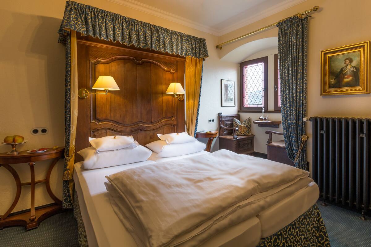 A hotel room with a large bed, two bedside lamps, a wooden headboard, a small table with a lamp, a picture frame, and a radiator.
