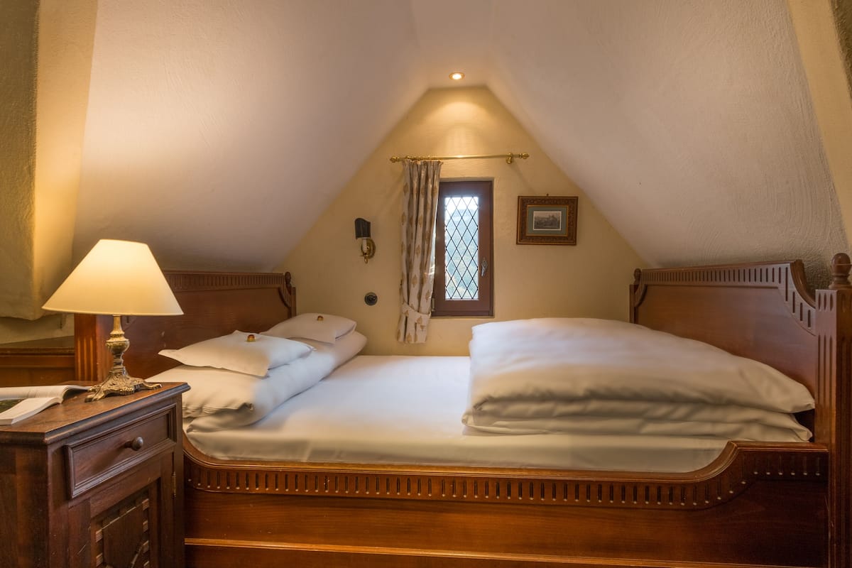 A single bed in a room with a sloped ceiling, a nightstand with a lamp, a window with curtains, and a framed picture on the wall.