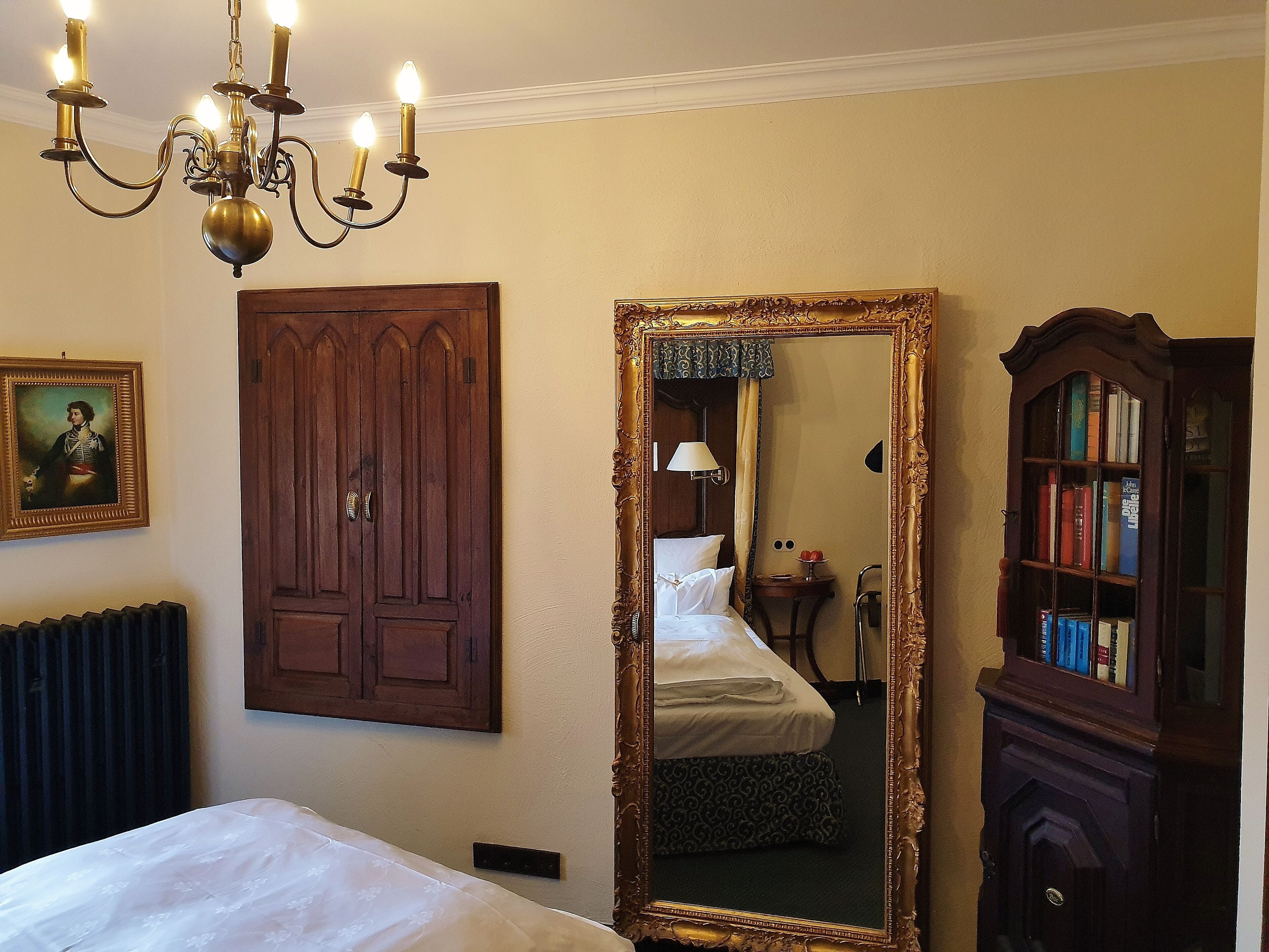 A hotel room with a large bed, a wooden wardrobe, a mirror, a chandelier, and a bookshelf.