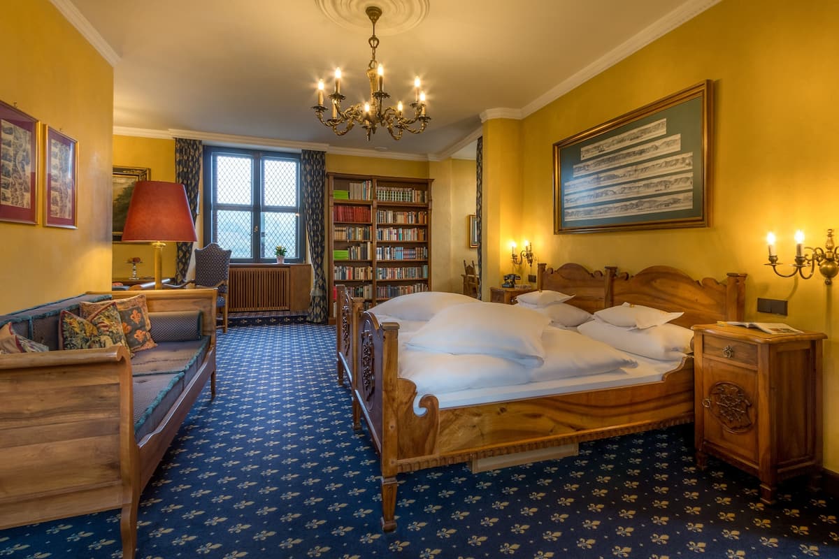 A room with a large bed, a wooden side table, a bookshelf, and a window with curtains.