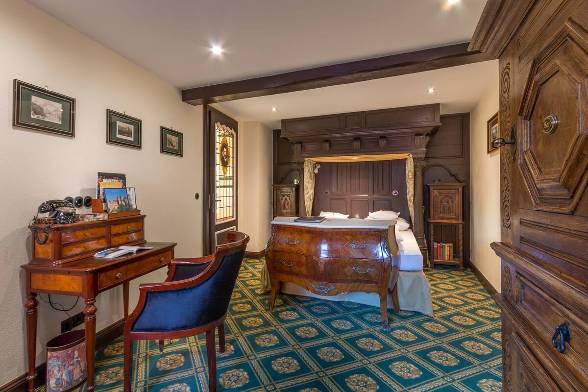 A bedroom with a wooden bed, a desk with a chair, a dresser, and a patterned carpet.
