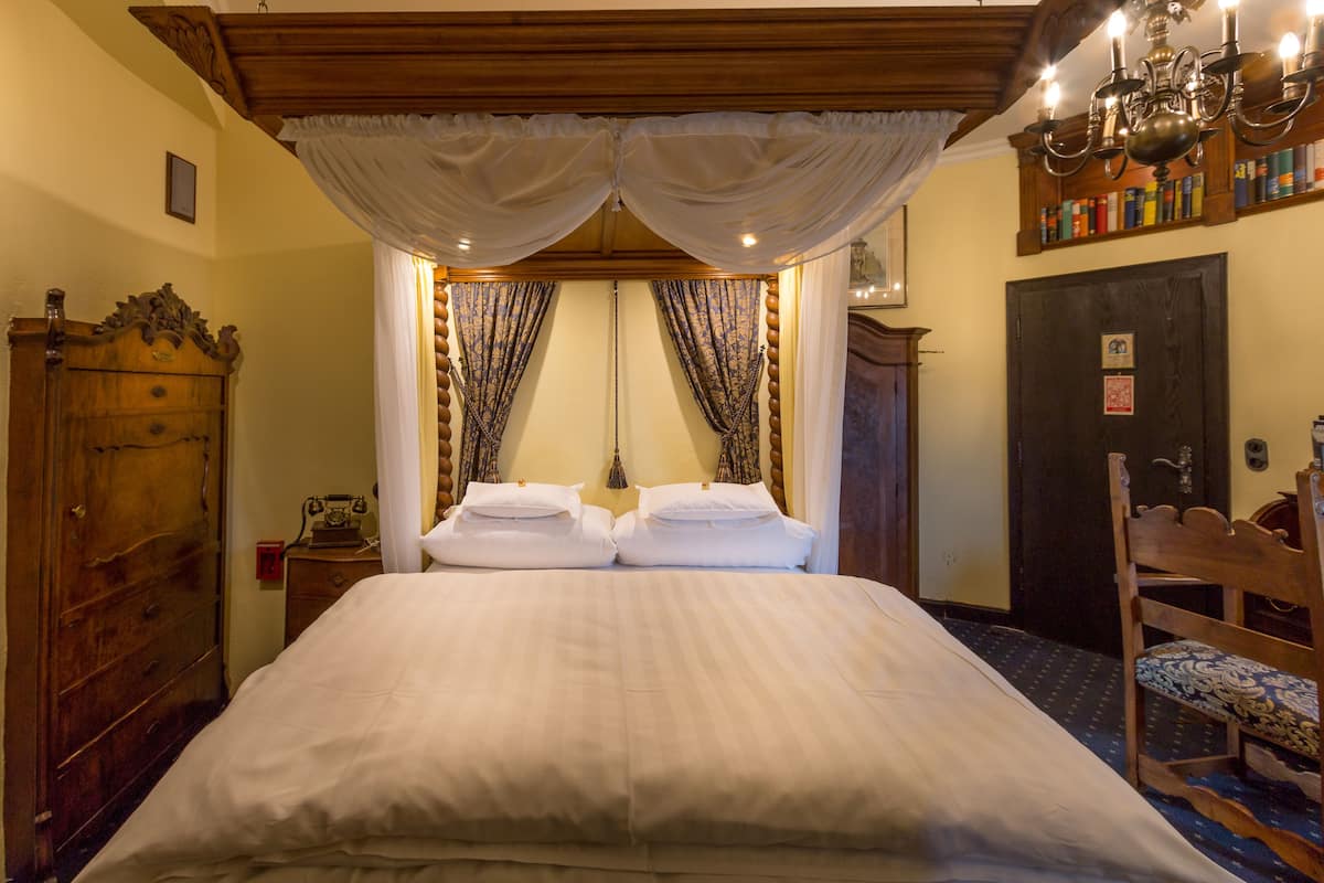A neatly made bed with a canopy, a wooden wardrobe, a chair with a patterned cushion, and a bookshelf with books.