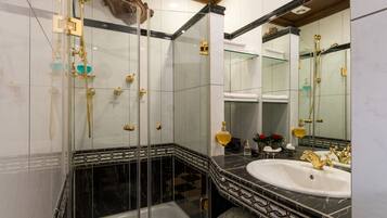 A luxurious bathroom with a glass-enclosed shower, a marble-tiled floor, a large mirror, and a white sink with gold fixtures.