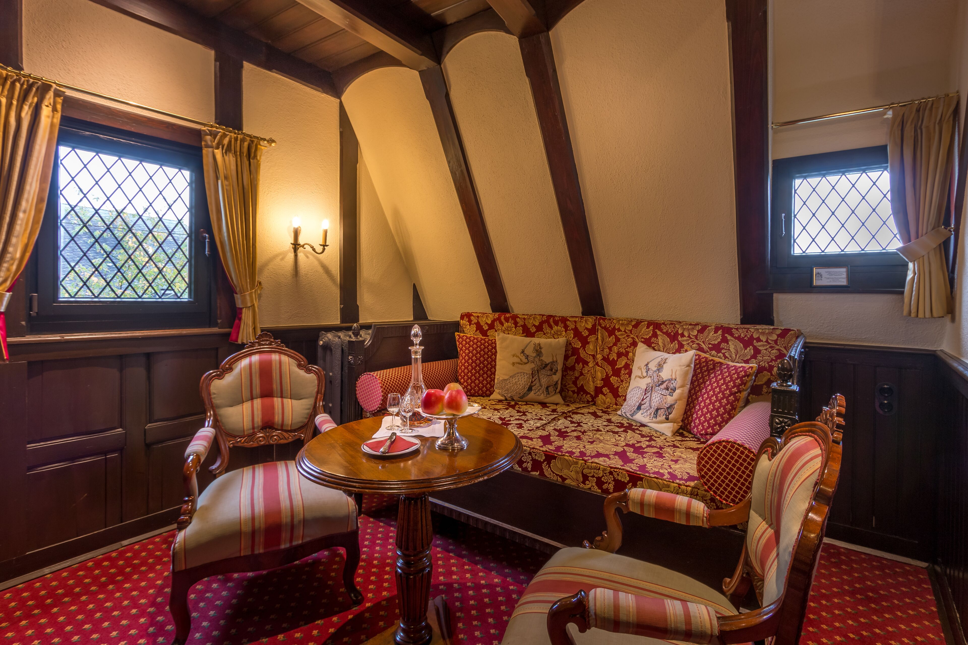 Ein gemütliches Zimmer mit Holzbalkendecke, einem gemusterten Sofa, gestreiften Sesseln und einem kleinen runden Tisch mit einer Vase und Obst.