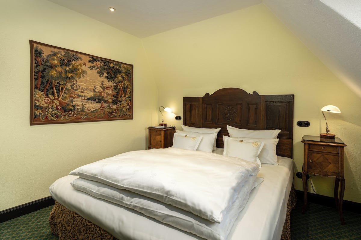 A neatly made bed with white linens, a wooden headboard, and a large landscape painting on the wall.