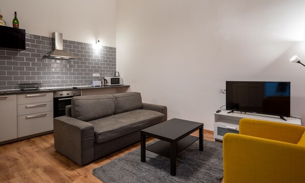 A modern living room with a grey sofa, a coffee table, a yellow armchair, a flat-screen TV, and a kitchen area with grey tiles and a microwave. - Lie On Me Lazy Simon (Budapest)