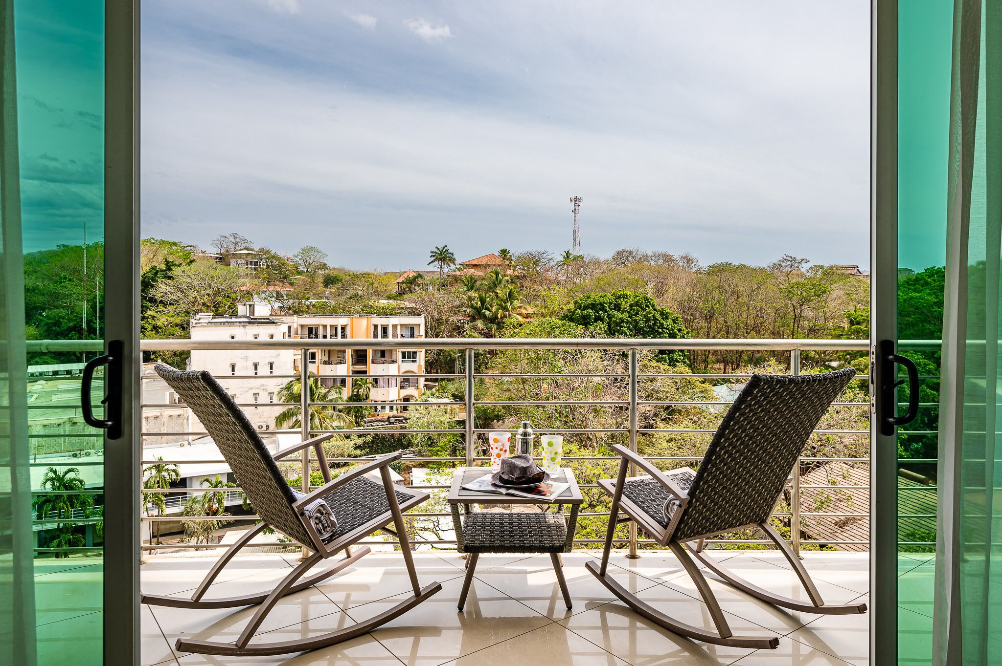 A balcony with two rocking chairs and a small table, overlooking a residential area with houses and greenery.