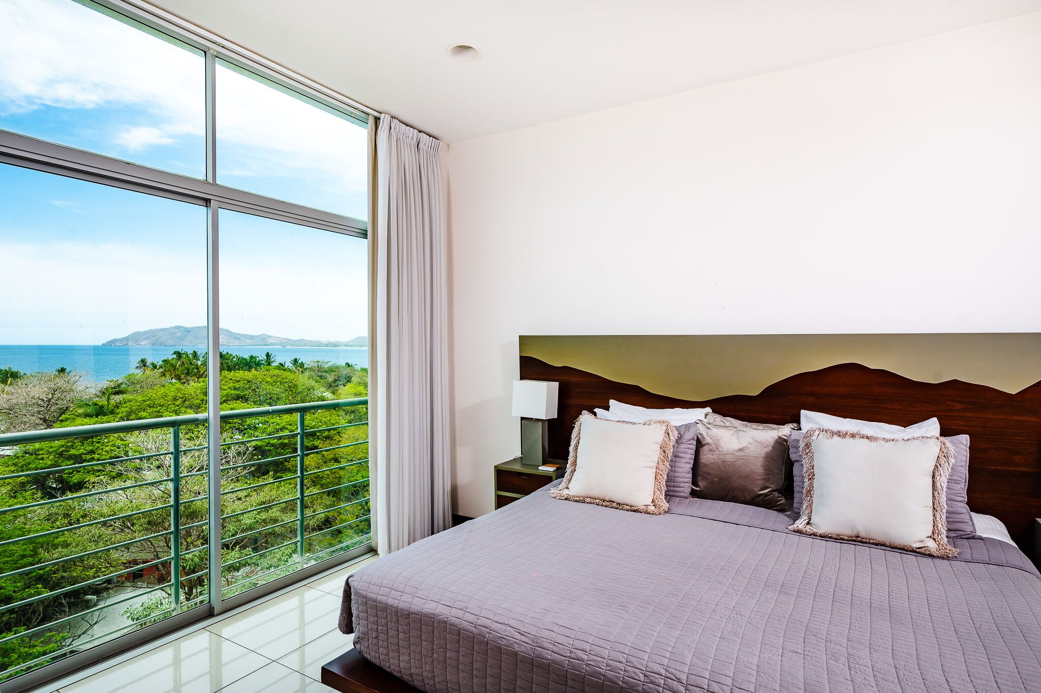 A bedroom with a large window, a bed with pillows, and a nightstand.