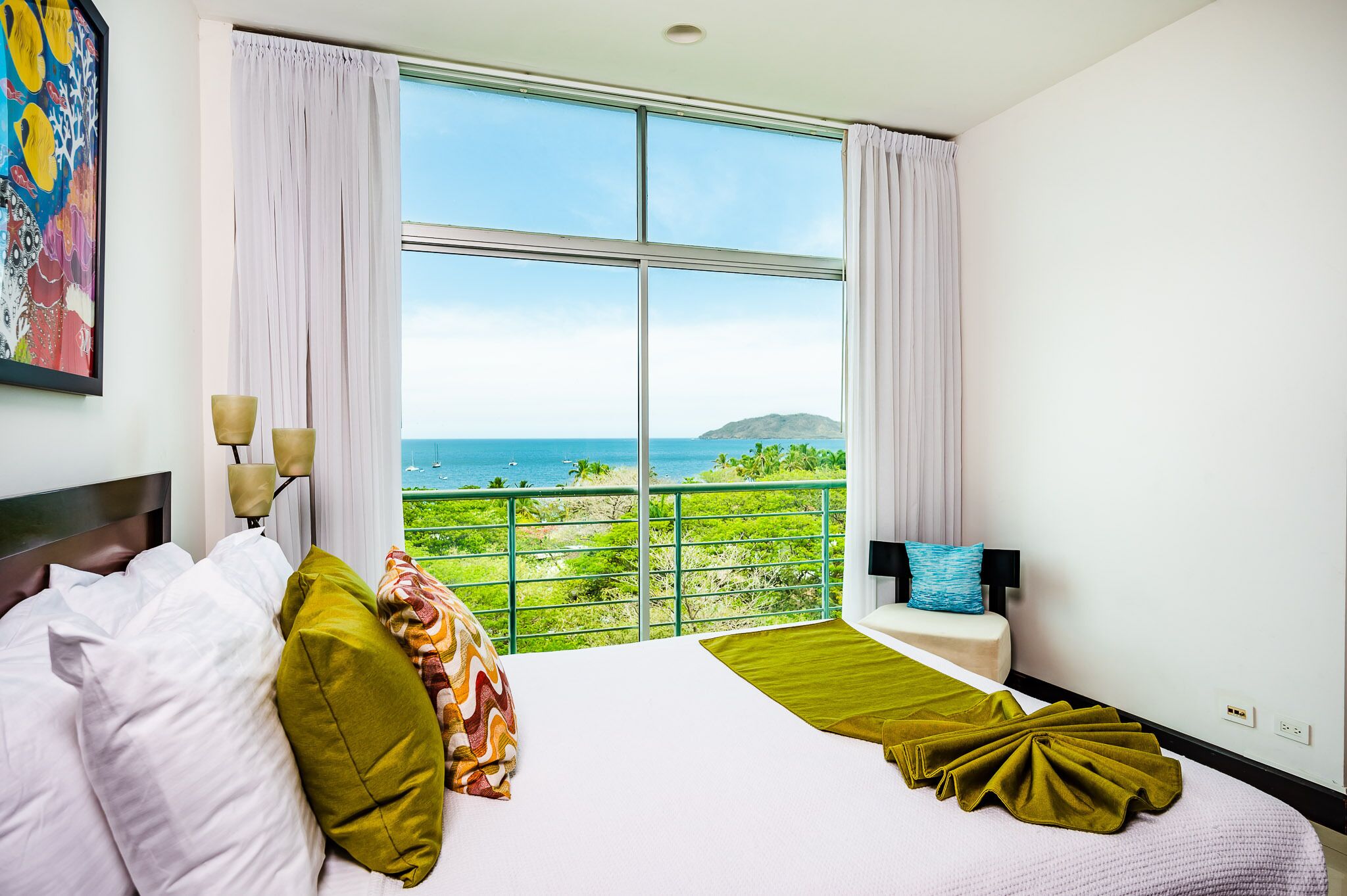 A hotel room with a large window, a bed with white and green bedding, a small bedside table with a lamp, and a colorful painting on the wall.