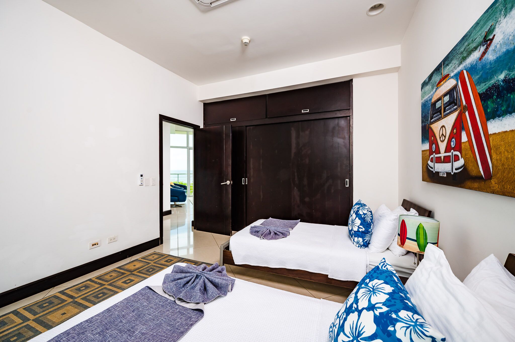 A hotel room with two beds, a large dark wardrobe, a painting of a van and surfboard, and a small bedside table with a lamp.