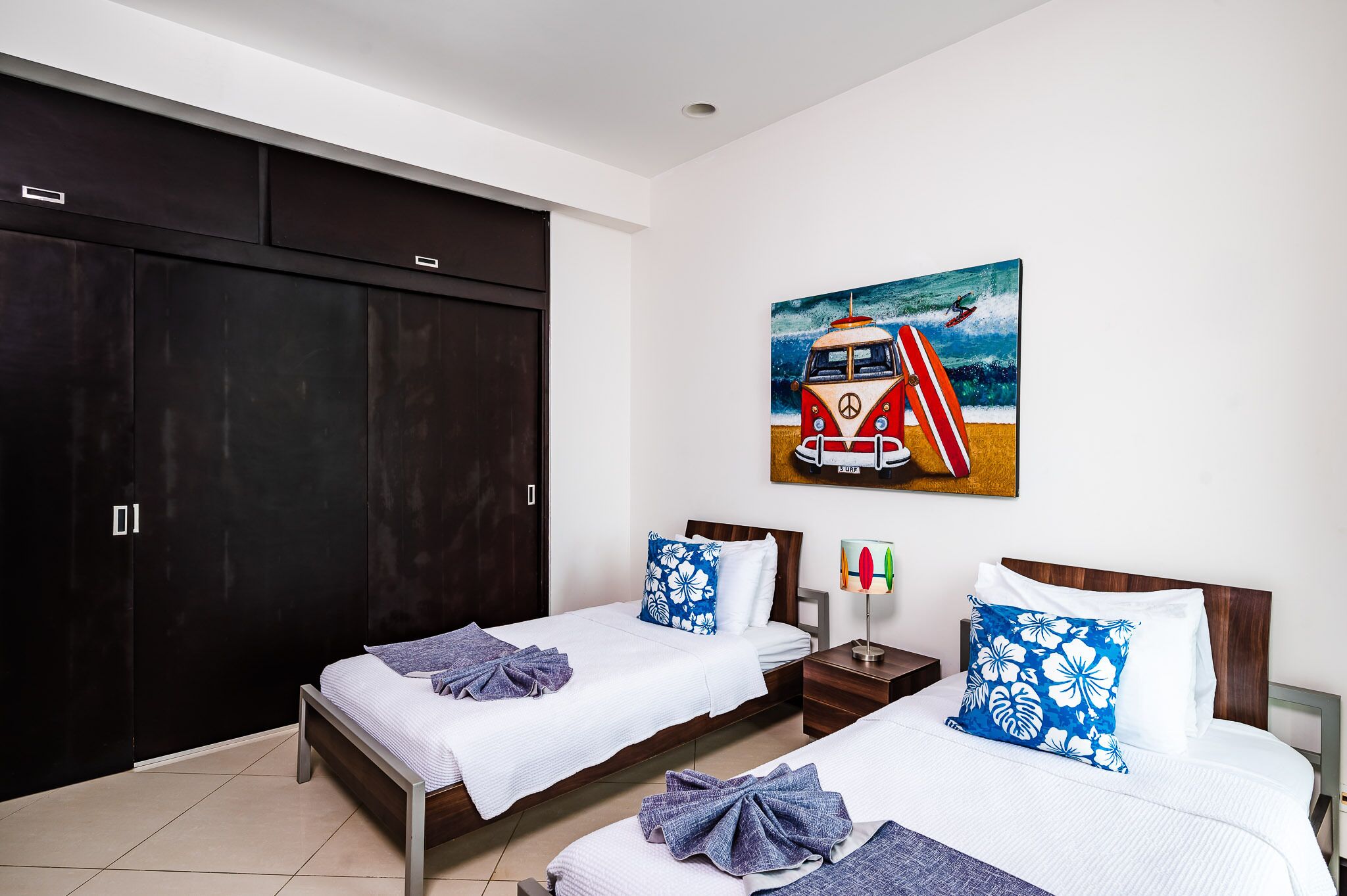 Habitación de hotel con dos camas, un armario oscuro, un cuadro de una furgoneta en la playa y una mesita de noche con una lámpara y una botella.