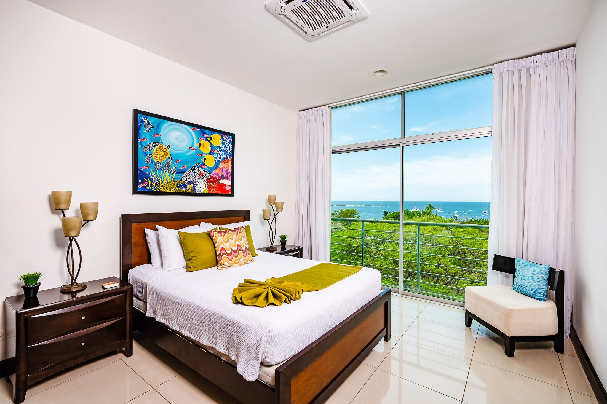A bedroom with a large bed, a nightstand, a chair, a painting, and a balcony with a view of the sea.