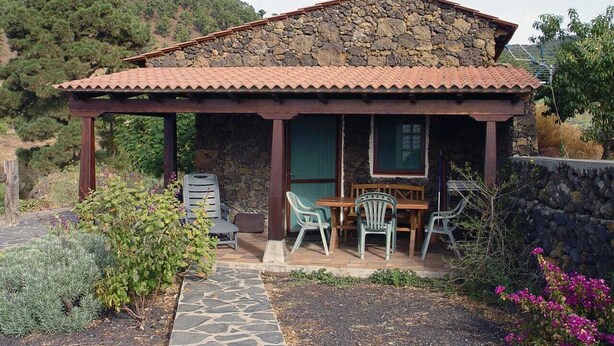 Ein Haus aus Stein mit einem Ziegeldach, einer überdachten Terrasse mit Tisch und Stühlen sowie einem Steinweg, der zum Eingang führt.