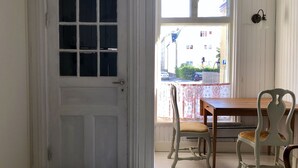 Ein Zimmer mit weißer Tür, einem Holztisch, zwei Stühlen und einem Fenster mit Blick auf die Straße.