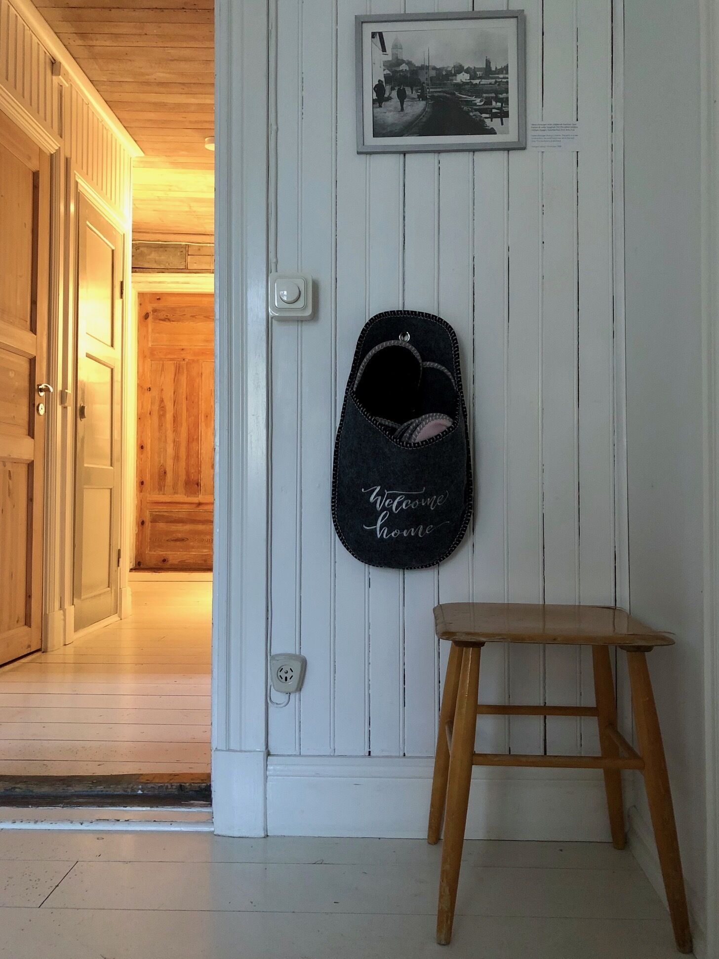 A hallway with a wooden door, a wall-mounted light switch, a framed picture, and a small wooden stool.