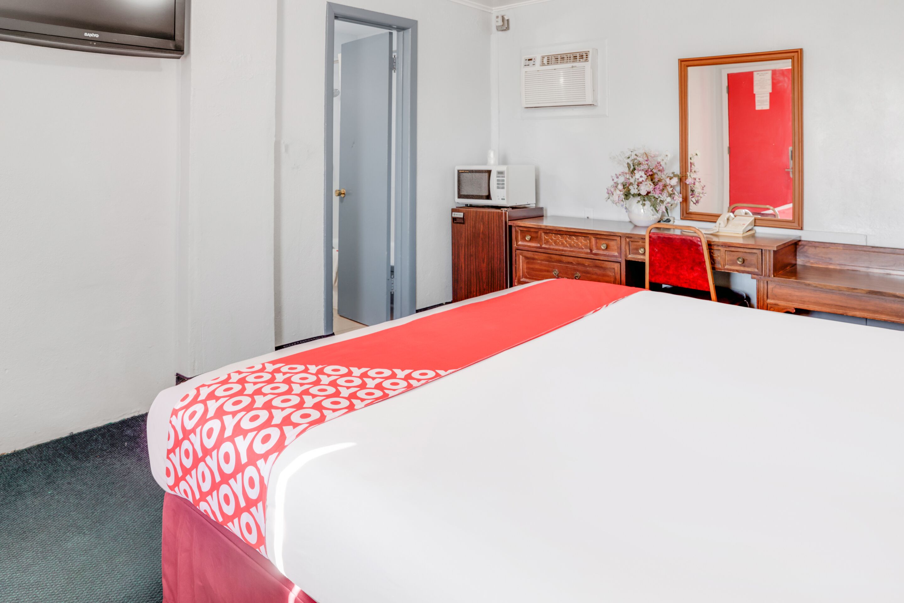 A hotel room with a bed, a desk, a chair, a dresser, a mirror, a microwave, and a door.