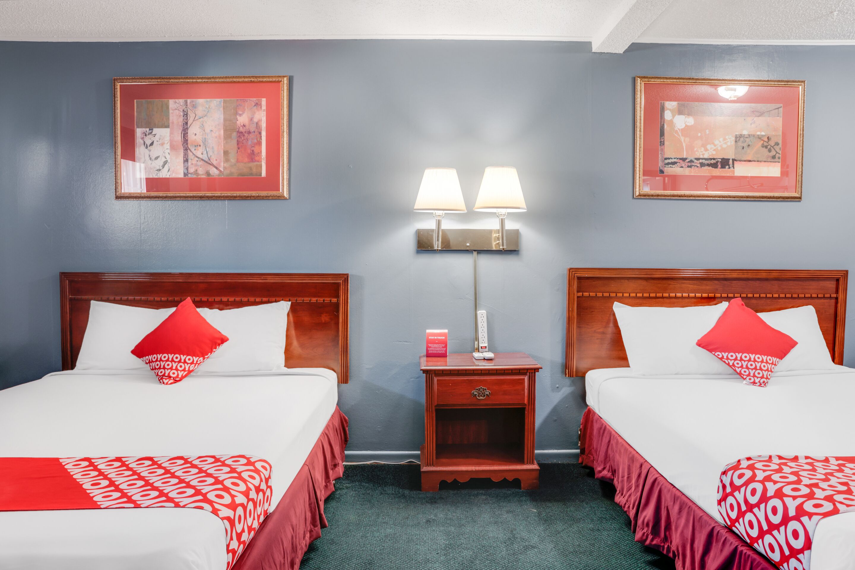 Quarto de hotel com duas camas, cada uma com colcha estampada em vermelho e branco, mesa de cabeceira com abajur e obras de arte emolduradas nas paredes.