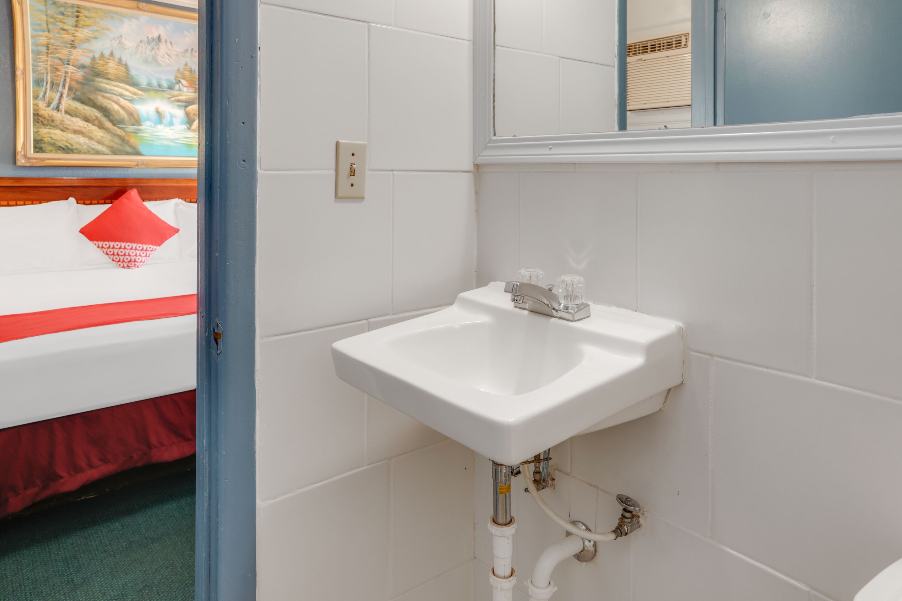 A bathroom with a white sink, a mirror above it, and a door leading to a bedroom with a bed and a red pillow.