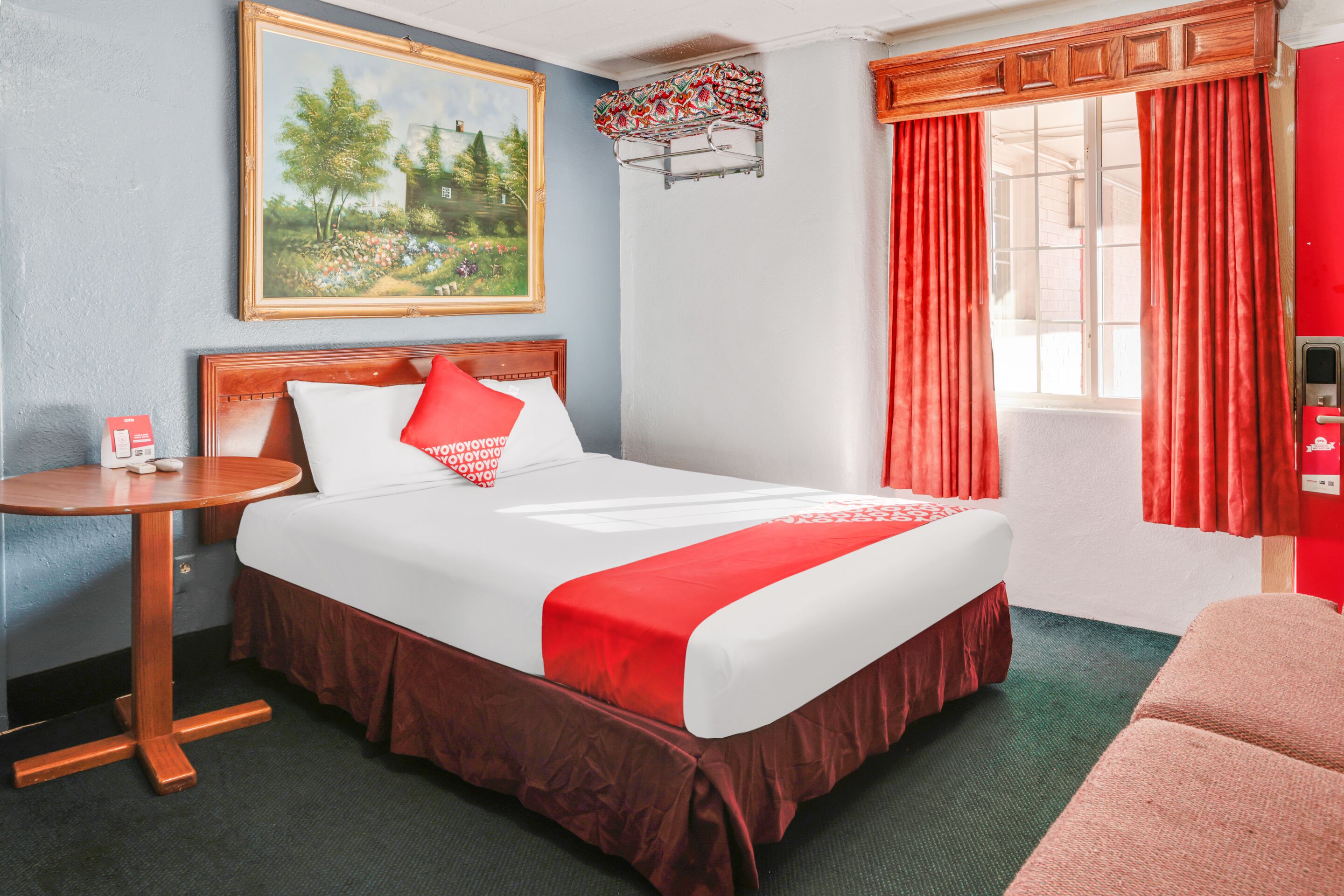 A hotel room with a bed, a small round table, a painting on the wall, and a window with curtains.