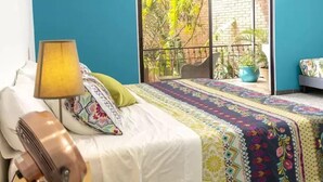 A bedroom with a bed, a guitar, a lamp, and a view of a brick wall and plants through a window. - Casa Orozco (Cali)