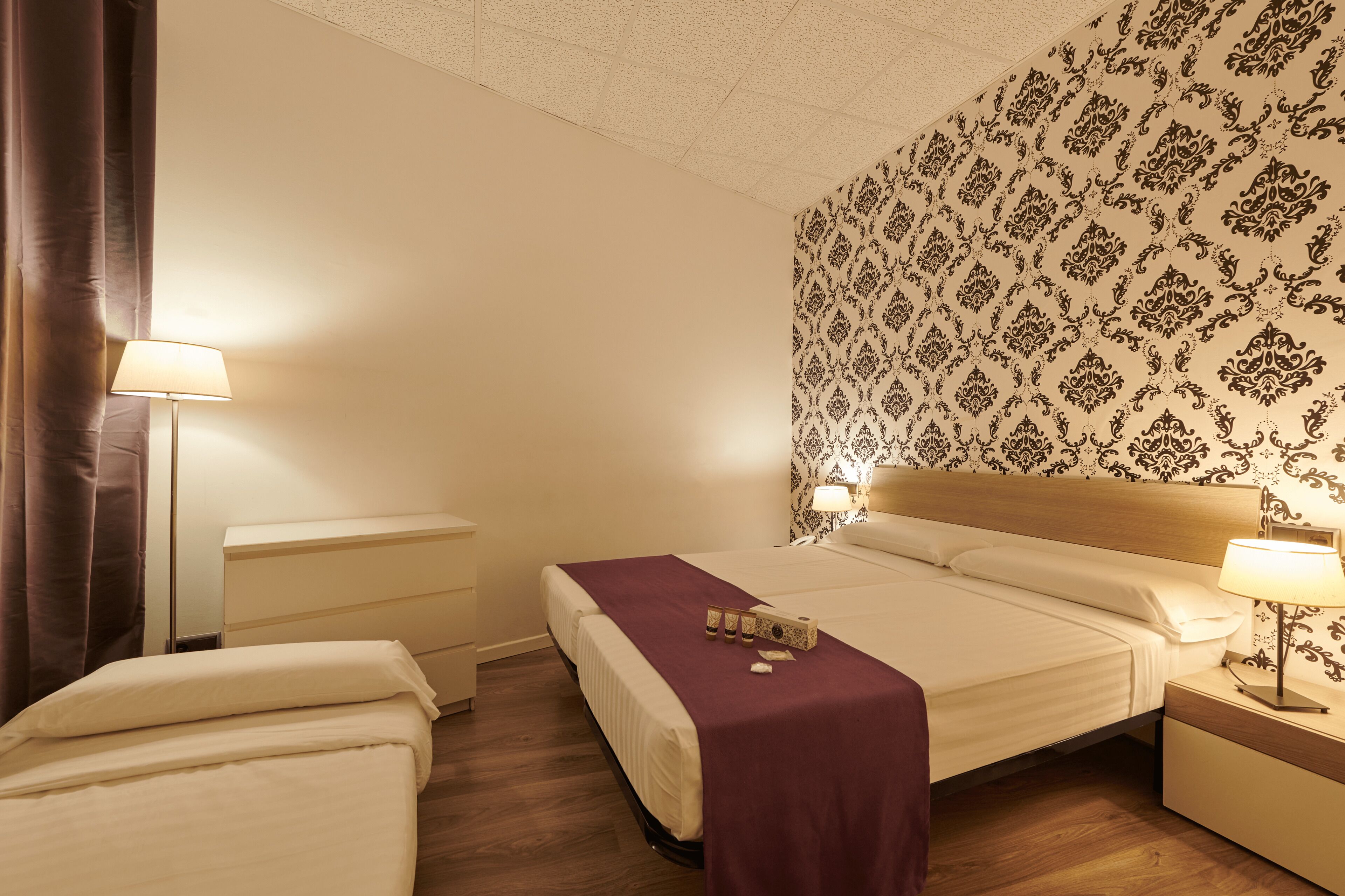 Hotellihuone, jossa on sänky, yöpöydät, lamppu ja kuviollinen tapetti.