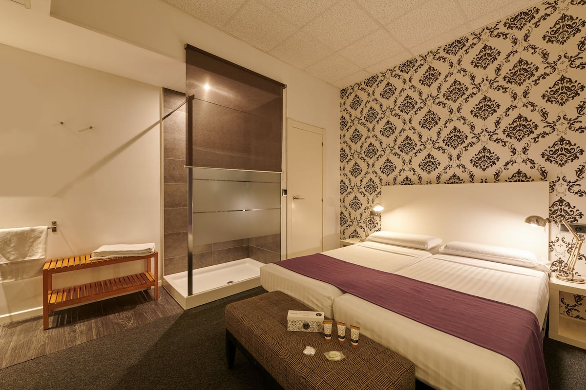 A modern hotel room with a large bed, a shower area, a wooden bench, and a patterned wall.