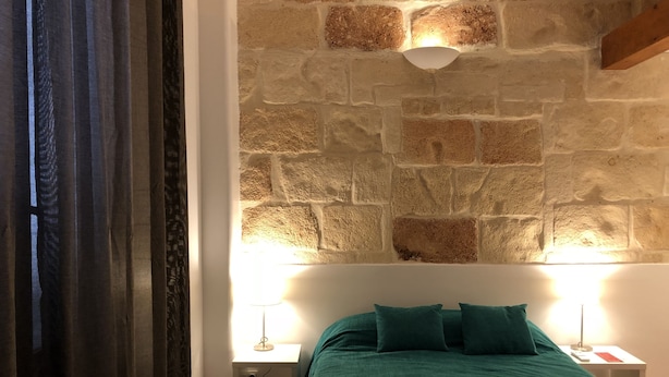 Een slaapkamer met een stenen muur, een groen bed, twee kussens, een nachtkastje met een lamp, en opgerolde handdoeken.