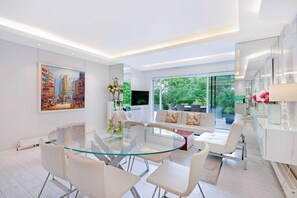 A modern interior with a glass dining table, white furniture, a large painting, and a view of greenery.