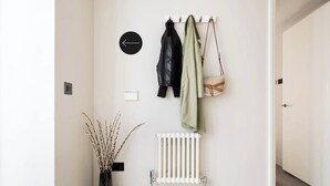 Un pasillo con un perchero lleno de chaquetas, un radiador, un reloj de pared y un florero con ramas.