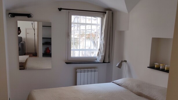 A bedroom with a large bed, a window with curtains, a radiator, a mirror, and a shelf with items. - Casa Daniele (010025-LT-1249) (Genova)