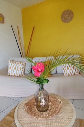 Une pièce avec un mur jaune, un canapé blanc orné de coussins à motifs, un vase en verre contenant une fleur rose et un set de table en fibres tressées posé sur une table ronde.