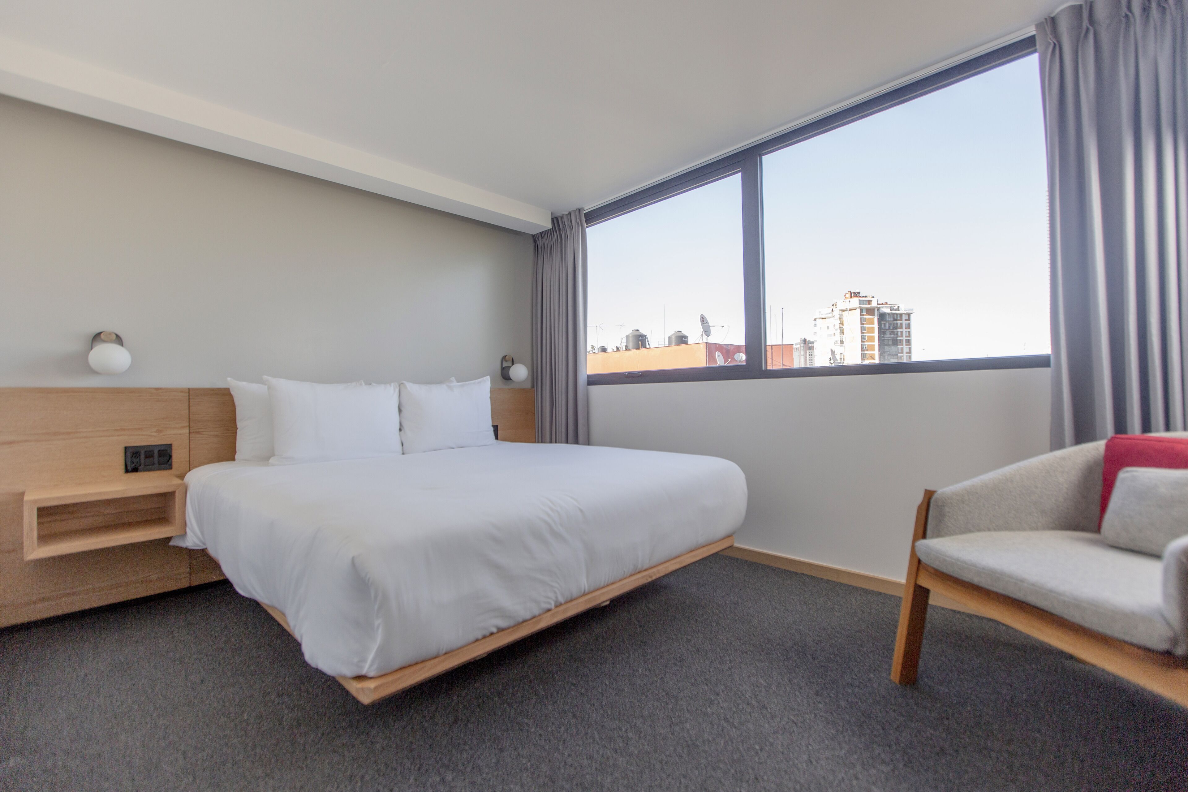 Ein Hotelzimmer mit einem großen Bett, einem Holfnachtkästchen, einem Sessel und einem Fenster mit Blick auf die Stadt.