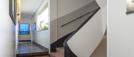 A modern interior with a polished floor, a staircase with a black handrail, and a hallway leading to a room with a window.