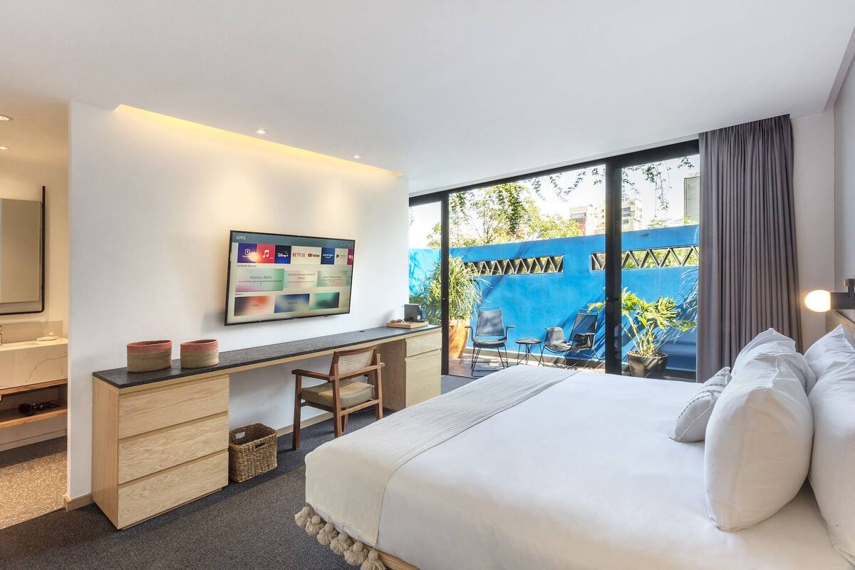 Une chambre d’hôtel moderne dotée d’un grand lit, d’un bureau avec un ordinateur, d’une télévision permettant d’accéder à des applications et d’un balcon offrant une vue sur la ville et la verdure.