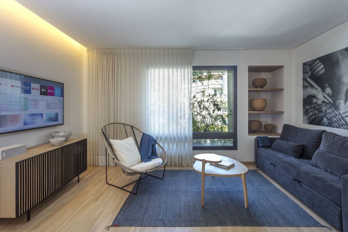 Un salon moderne avec un canapé gris, un fauteuil en osier, une petite table ronde, un téléviseur fixé au mur et une fenêtre donnant sur un paysage verdoyant.