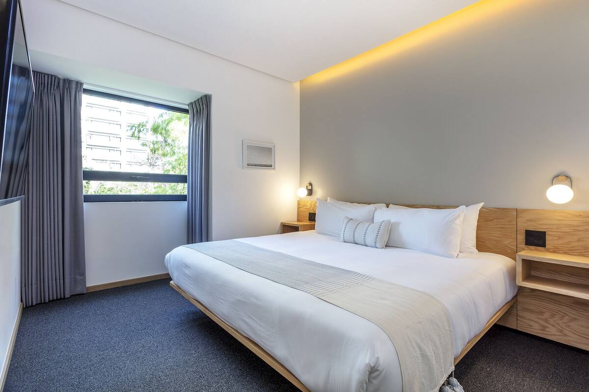 Une chambre d’hôtel avec un grand lit, deux tables de chevet, une fenêtre avec des rideaux et un luminaire fixé au mur.