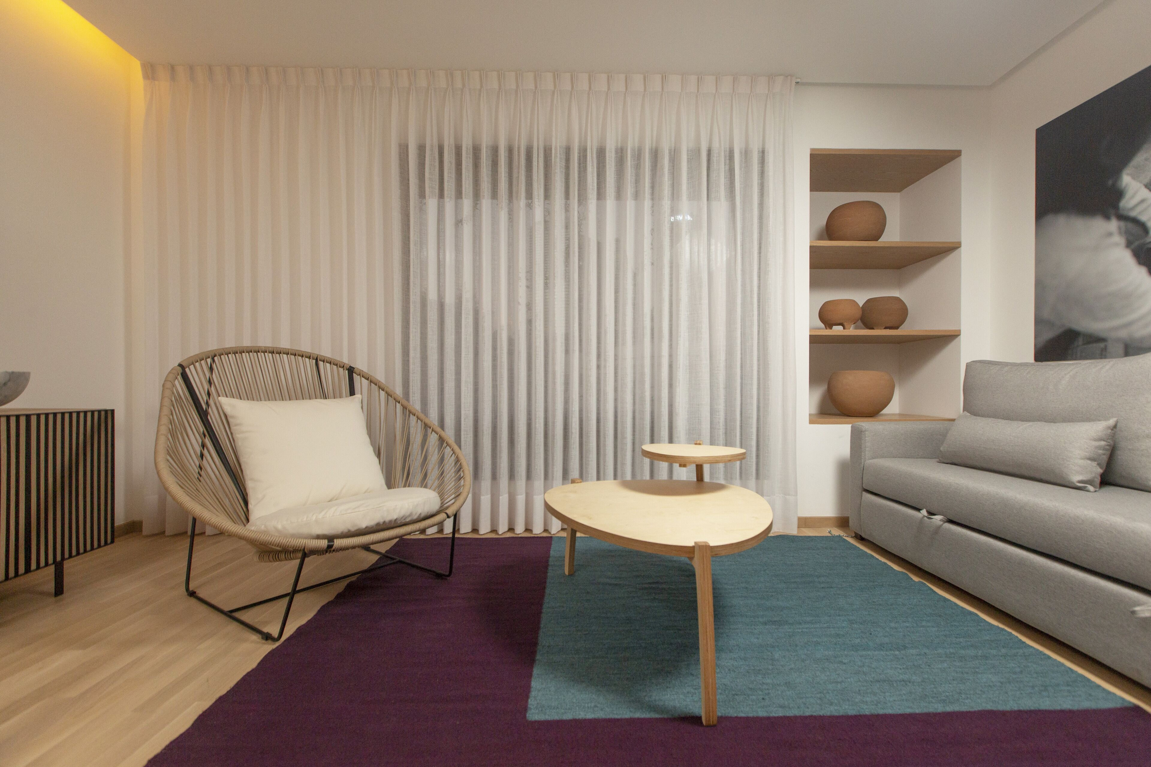 A modern living room with a wicker chair, a grey sofa, a small wooden table, and a decorative shelf with vases.