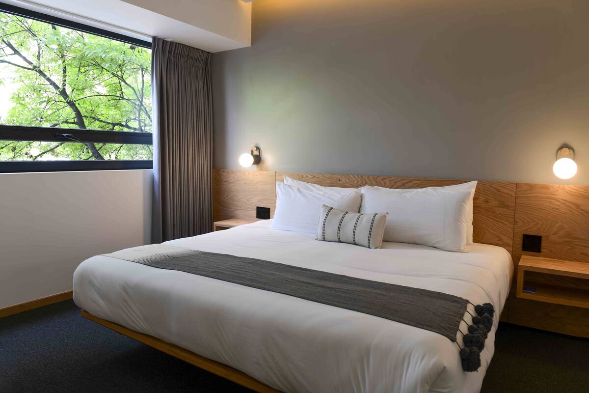 Une chambre d’hôtel moderne avec un grand lit, une tête de lit en bois et une fenêtre avec des rideaux.