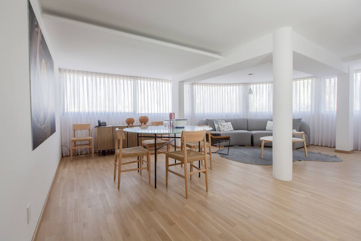 Un vaste espace de vie comprenant une table ronde en verre entourée de chaises, un canapé gris et une grande colonne blanche.
