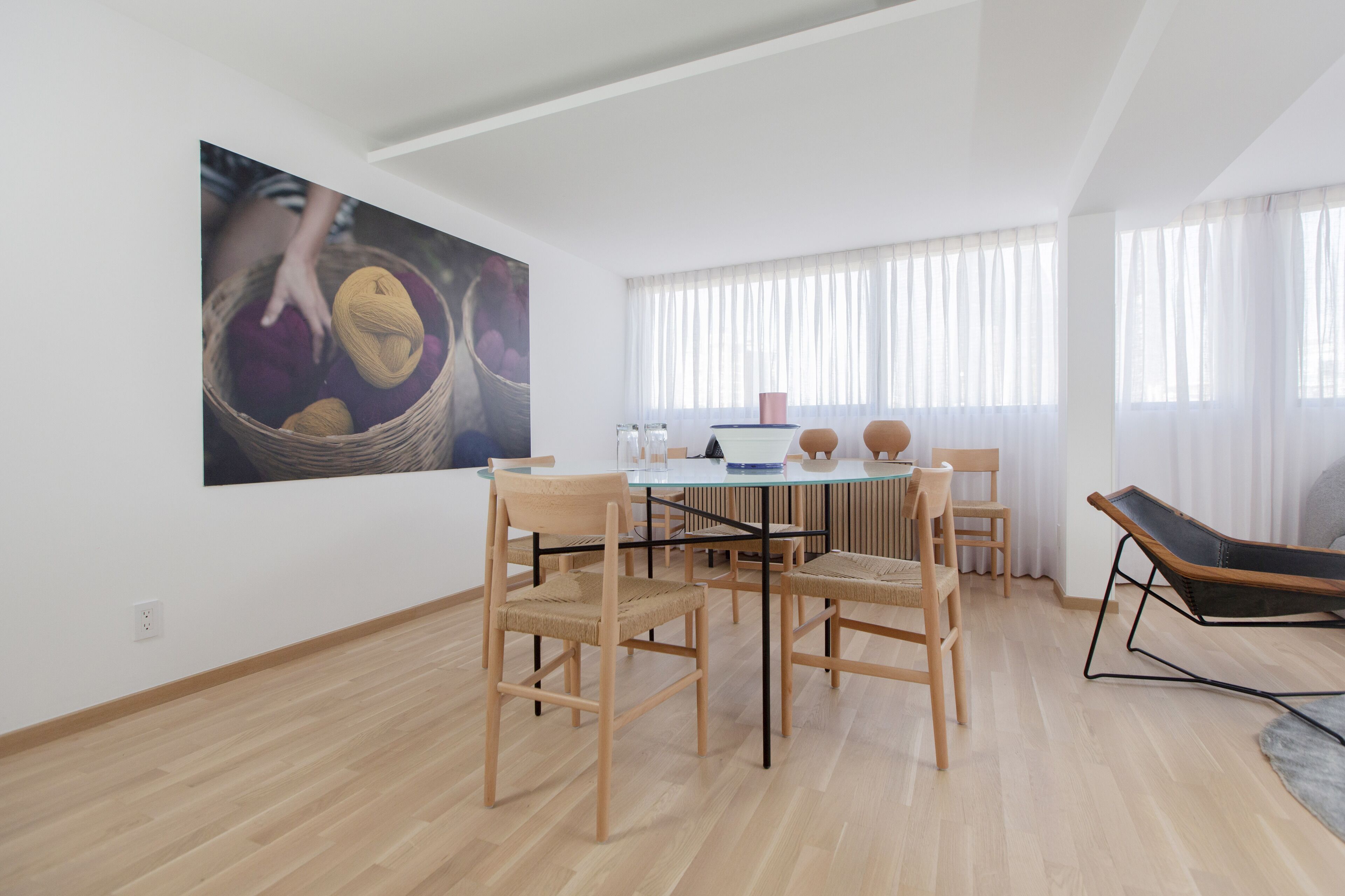 A modern living room with a glass-top dining table surrounded by wooden chairs, a large abstract painting on the wall, and a large window with sheer curtains.