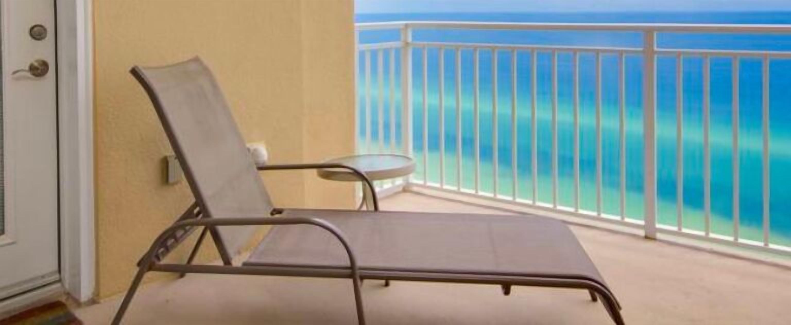Ein Balkon mit einem Liegestuhl und einem kleinen Tisch, mit Blick auf das klare, blaue Meer.