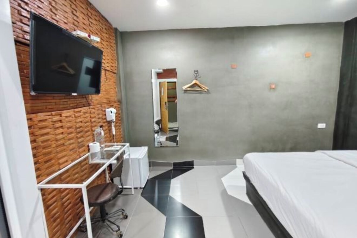 A hotel room with a TV mounted on a brick wall, a desk with a chair, a bed, a mirror, and a coat hanger.