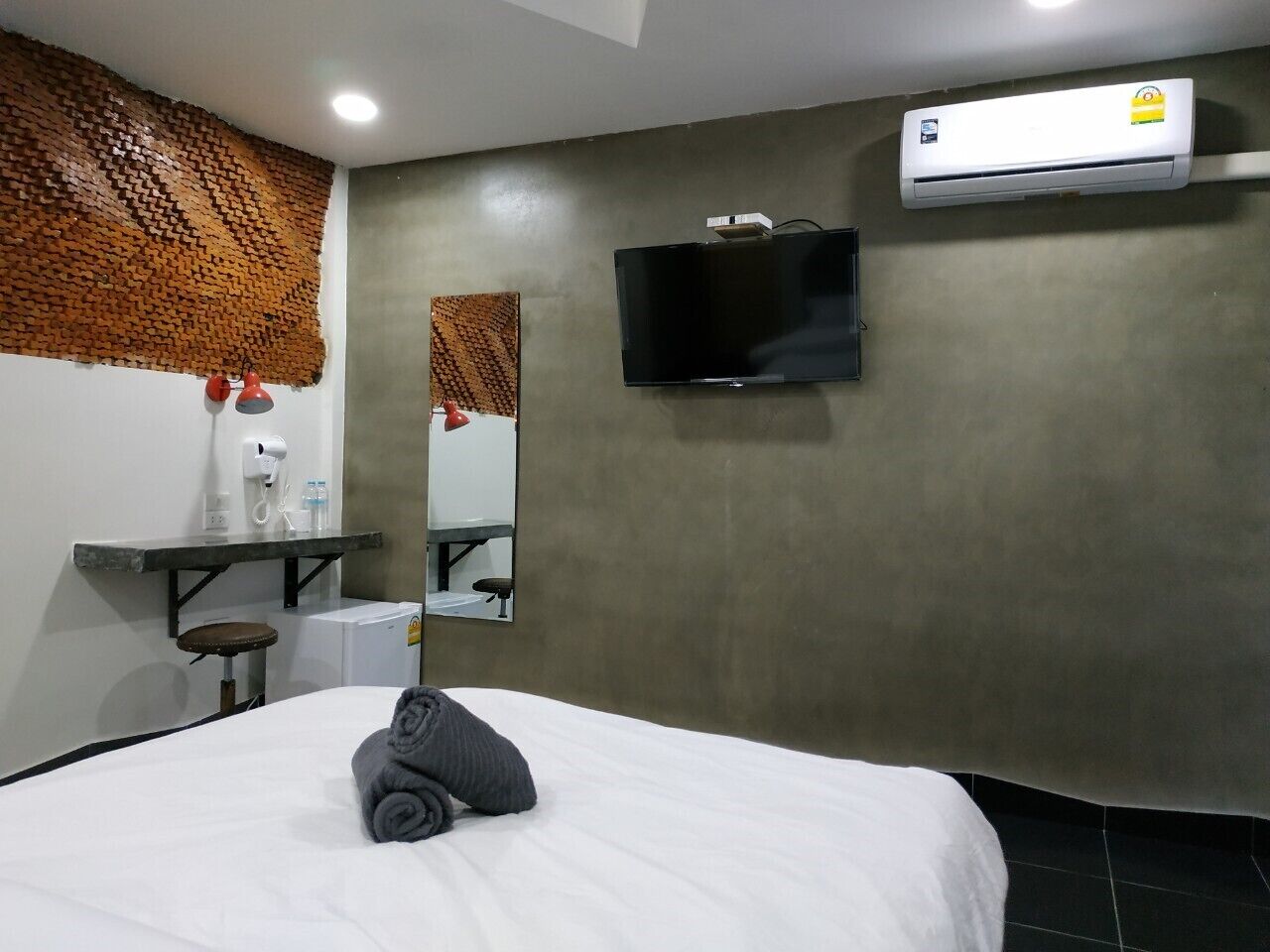A hotel room with a bed, a flat-screen TV mounted on the wall, a built-in air conditioning unit, a small stool, and a mirror above a vanity area.