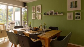 Un comedor con una mesa de madera preparada para cuatro personas, rodeada de sillas. Hay cuadros enmarcados y una pequeña estantería con objetos decorativos en la pared.