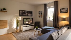 Una sala de estar moderna con un televisor de pantalla plana, un sofá, una mesa de centro y una ventana con cortinas.