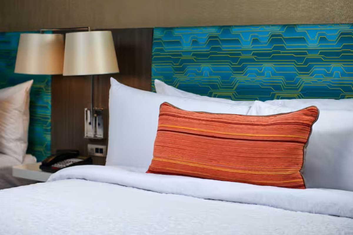 A hotel room with a bed, pillows, a nightstand, a lamp, and a patterned wall.