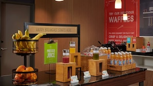 A breakfast buffet area with a sign that reads "HAPPY BREAKFAST STARTS HERE." There are bowls of fruit, a juice dispenser, and a display of orange juice bottles. - Hampton Inn Sherwood Portland (Sherwood)