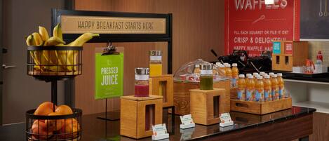 A breakfast buffet area with a sign that reads "HAPPY BREAKFAST STARTS HERE." There are bowls of fruit, a juice dispenser, and a display of orange juice bottles.